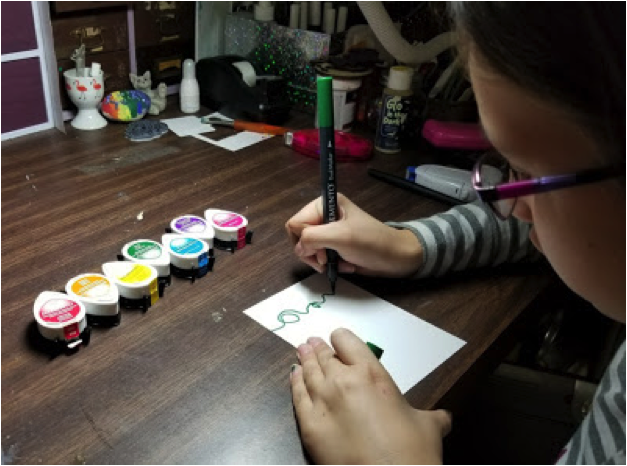 A girl draws a squiggly green line across a piece of white cardstock to represent a string for Christmas lights.